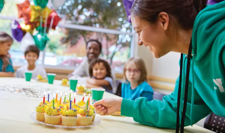 Photo shows YMCA birthday party with woman lighting cupcake candles